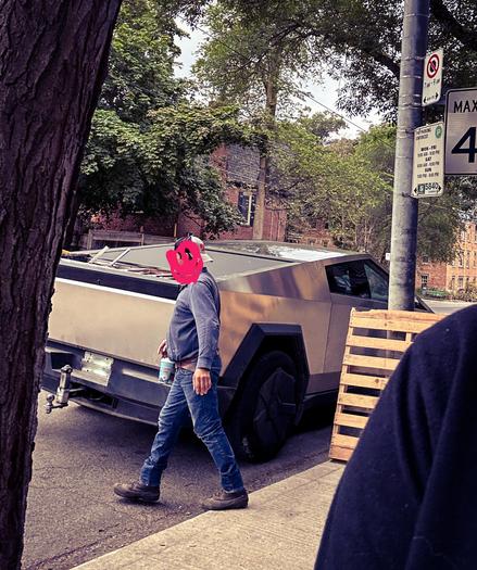 A man walking behind a cybertruck (face obscured). The rolltop of the trunk is partially open with scrap wood sticking out of it. There is a wooden pallet propped up against a pole. There is no hope of getting it in the back.