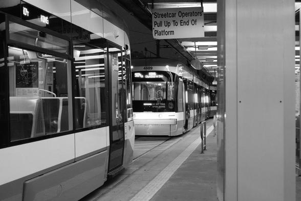 The outdoor platform. At right are two streetcars waiting, receding to a vanishing point at the far end of the platform. The one closest to the camera fills the frame, top to bottom at the left side of the image. The car in the distance has it's doors open. Above them, a sign reads "Streetcar operators pull up to the end of the platform" At far right of the image, running top to bottom, is the plain rectangle of a support pillar for the roof.