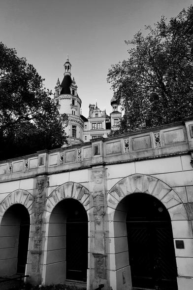 A low-angle, black and white photograph of Schwerin Castle in Germany, peeking over a textured stone wall with three prominent arched alcoves. The castle's multiple ornate turrets and detailed architecture are visible, framed by dark trees on either side against a lighter sky. The image conveys a sense of historical grandeur and architectural beauty.
