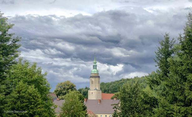 Widok na wieżę kościoła z zieloną wieżyczką, otoczoną drzewami i dachami pod zachmurzonym niebem.