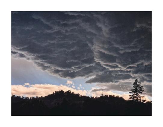 This photo is of a dramatic, dark sky filled with heavy, grey clouds that take up the upper two-thirds of the frame.  These clouds have a textured appearances. Below, silhouetted against the clouds and a piece of lighter sky, is a line of dark hills or mountains, sparsely dotted with trees. A tall evergreen tree is visible on the right and it stands out against the dark sky.