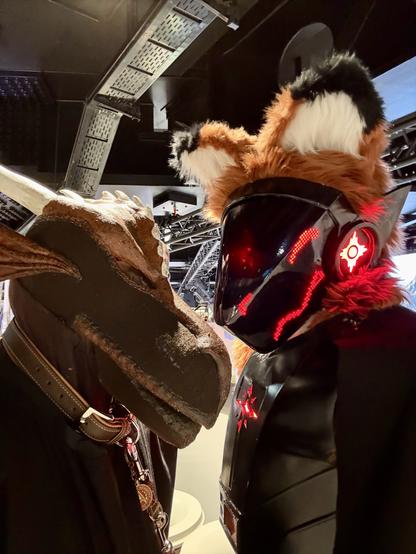 A dragon-themed costume features a character with a detailed head and collar, while a second character in a futuristic, armored suit with glowing elements and a fox-like head pose closely together in a modern setting.