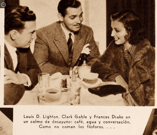 Tres personas sentadas en una mesa de desayuno: un hombre y una mujer conversan con un tercer hombre, todos vestidos elegantemente, en un ambiente acogedor.