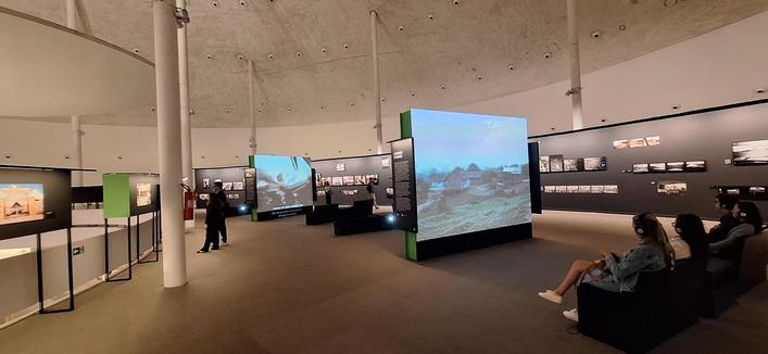 Mezanino da galeria principal, onde acontece a exposição. Projetores mostram filmes do autor, com pessoas sentadas em poltronas assistindo. Ao fundo fotos dispostas em uma parede preta. É possível ver parte da abóboda do teto interno do museu, que cobre o espaço