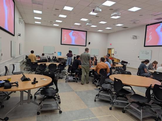 A large room with fluorescent lighting. Three large screens, one high on each wall. They show a purple and blue overhead shot of a 3D videogame river. Below, at circular tables, groups of people play original games
