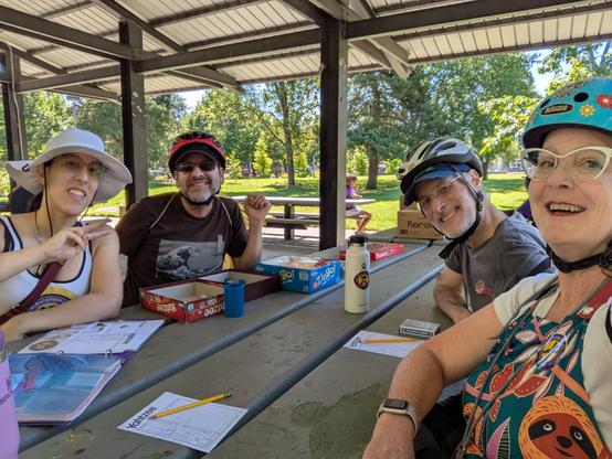 4 people sitting around a park bench table under roof playing Yahtzee! wearing bike helmets. Alliana wearing a Genital Autonomy Collective tank top.