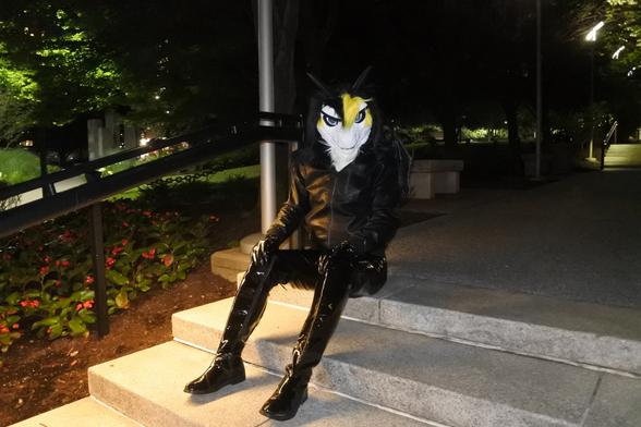 Fursuit photo: Night-time shot of Terry the dragon in a small park behind the Omni hotel in Pittsburgh, wearing PVC jeans and a leather jacket.  He has a sinister grin