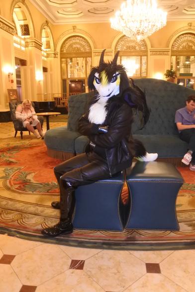 A dragon fursuiter in leather jeans sits on a couch in the lobby of the Omni hotel