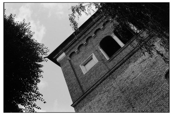 The image features a brick tower viewed from a low angle, creating a sense of height and grandeur. The tower is constructed from red bricks, with a rough texture visible on the surface. It has a series of arched windows, with one arched window being open, revealing a dark interior. Above the windows, there is a decorative cornice with a scalloped edge. The tower is partially obscured by tree branches, which add a natural element to the scene. The sky is visible in the background, with a few clouds scattered across it. The image is in black and white, which emphasizes the textures and contrasts within the scene.