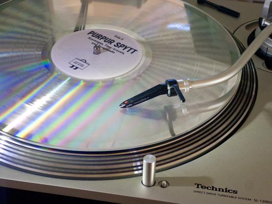 A silver disc spinning on a record player. Tell-tale rainbow refractions give it away as a LaserDisc, but the needle is clearly tracking grooves. Purpur Spytt,, reads the plain white label.