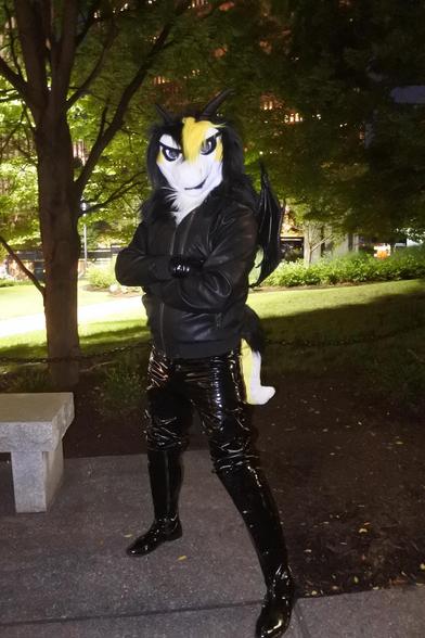 Fursuit photo of Terry the dragon in a small area of parkland behind the Omni hotel in Pittsburgh.  It is night, and the dragon's PVC gloves and jeans reflect the camera flash and streetlights

Photo by Aisha DeCabre