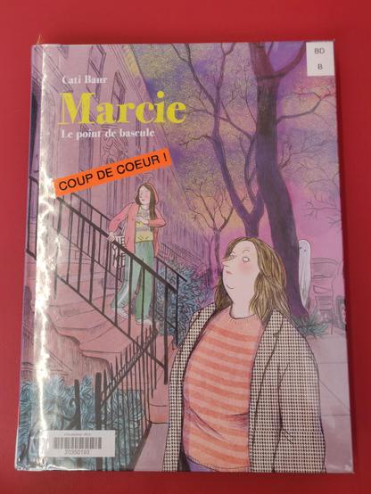 Couverture de la BD avec un bandeau "coup de cœur" apposé par la mediatheque. La BD s'appelle Marcie, le point de bascule, de cati Baur. On y voit au 1er plan l'héroïne de 50 ans qui est sur un trottoir et derrière une jeune fille descendant les escaliers du perron d'un immeuble. Dans le fond, caché derrière un arbre, une personne déguisée en fantôme.