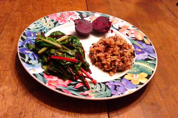 A plate of food on a wooden table. The plate is white and decorated with leaves and flowers against a black band around the edge. In the plate are two halves of a purple-red beet, some colorful cooked greens, and multi-colored rice.