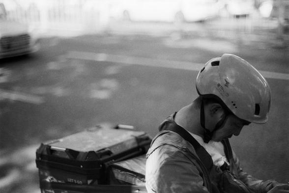 Fujifilm Neopan 100 SS (FF)

**English:**
A close-up black-and-white photograph of a delivery person wearing a helmet and riding a bicycle. The cyclist is looking down, possibly checking something on the bike. The background is blurred, emphasizing the focus on the cyclist and the delivery box attached to the back of the bicycle.

**Chinese:**
这张黑白照片特写了一位戴着头盔的送货员骑自行车。骑车人低头看着，可能在检查自行车上的某些东西。背景是模糊的，强调了对骑车人及自行车后面所附送货箱的关注。