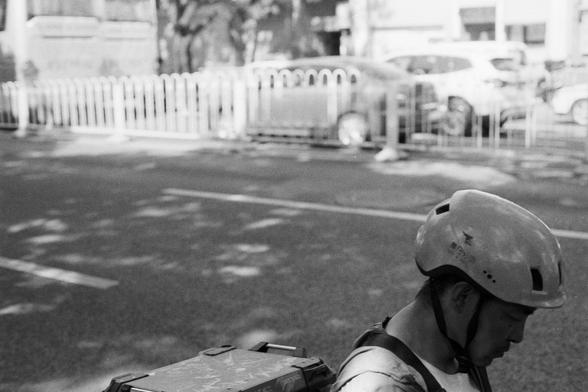 Fujifilm Neopan 100 SS (FF)

**English:**
A black-and-white photograph captures a delivery person wearing a helmet and riding a bicycle on a city street. The cyclist is carrying a large box on the back of the bike. The background shows a slightly blurred urban scene with a sidewalk barrier and some vehicles, indicating motion and the hustle of city life.

**Chinese:**
这张黑白照片拍摄了一位戴着头盔的送货员在城市街道上骑自行车。骑车人在自行车后面携带着一个大箱子。背景展示了一个略微模糊的城市场景，有行人护栏和一些车辆，显示出运动和城市生活的忙碌。