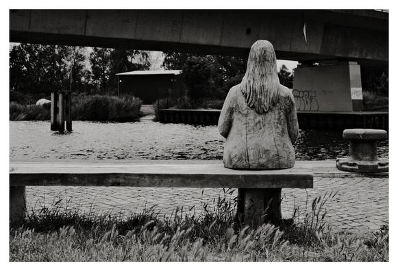 Schwarz-Weiß-Aufnahme einer Holzskulptur einer Person mit langen Haaren, die auf einer Bank am Wasser sitzt. Im Hintergrund verläuft eine Betonbrücke über den Fluss oder Kanal. Das Ufer ist bewachsen, und auf einem Brückenpfeiler ist Graffiti zu sehen.