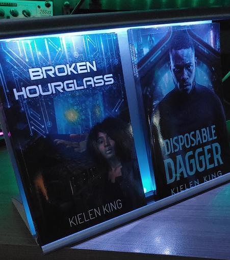 Two books inside of an illuminated book stand. Left: Broken Hourglass. Right: Disposable Dagger.