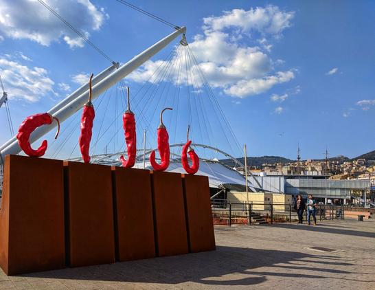 A sunny outdoor shot shows five large, glossy red chilli sculptures, each with a curled stem, resting on a dark, reddish-brown rectangular base. In the background, behind the sculptures, a large white mast and rigging from a sailing ship are visible, along with parts of the harbour in Genoa. The sky above is bright blue with scattered white clouds. To the right, a paved area extends where two people are walking, and further back, harbour buildings and a hillside are visible.