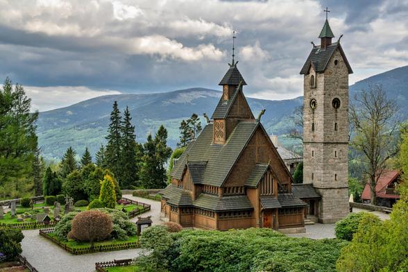 Im Vordergrund ein gepflegter Garten und mitten auf dem Bild die Stabkirche Wang. Im Hintergrund Berge. Das Riesengebirge.