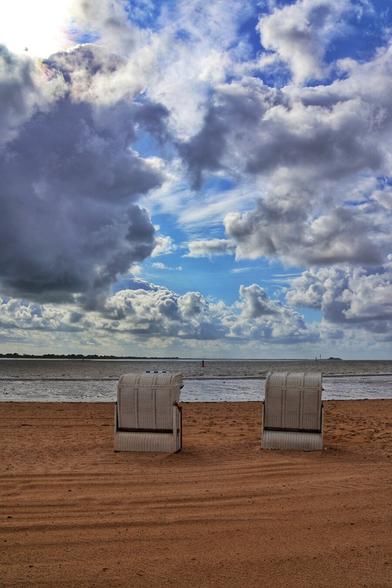 A slightly high-angle, full shot of a beach with two white, wicker beach chairs (Strandkörbe) facing the sea under a dramatic sky. The chairs are positioned in the foreground on the orange-brown sand, with their backs to the viewer. The sea, appearing choppy, stretches across the mid-ground. The horizon features a faint line of land and what appears to be a red buoy. The sky is a mix of bright blue with fluffy white clouds towards the centre, contrasting with dark, heavy grey clouds on the left and right, particularly overhead. A sunbeam breaks through the clouds on the top left.