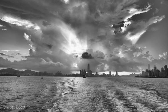 This is a black and white cityscape photo with an expanse of water in the foreground and a developing tropical storm overhead A city. The lower quarter of the image is filled with a scene of dark harbour water. In the distance, about 1km away is a city made up of multiple high-rise buildings. To the left is a ragged ridge of high terrain. In the centre is a large tropical thunder storm (cumulonimbus) with a low flat cloud base and stretching horizontally about 5-10km across the city and beyond. The cloud develops vertically with the centre being bright from the low rising sun that's behind. At a point about three-quarters up in the image the cloud spreads horizontally more to the left than right, possibly forming an  'Anvil' that tops a classic thunderstorm. To the left and right of the storm is a near clear and cloudless dark sky.
The location: The body of water is Victoria Harbour. The city is Hong Kong (2018).