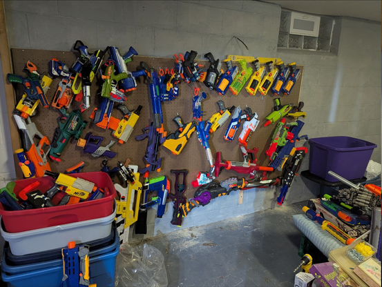 A photo of a grim basement wall with four large pegboards mounted to it, covered in colorful Nerf blasters of all shapes and sizes.
