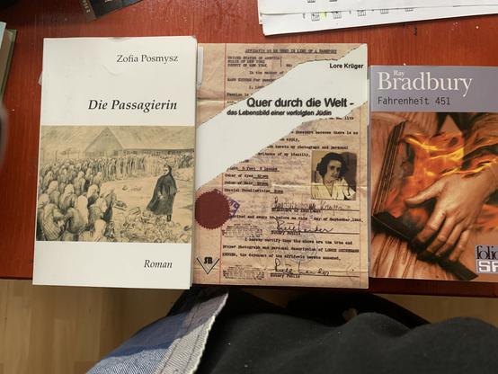 Das Bild zeigt drei Bücher auf einem rotenTisch. "Die Passagierin" von Zofia Posmysz, "Quer durch die Welt" von Lore Krüger und "Fahrenheit 451" von Ray Bradbury.