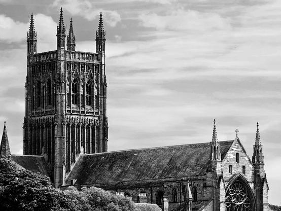 A dramatic black and white photograph of Worcester Cathedral in England, showcasing its impressive Gothic architecture under a cloudy sky. The tall, ornate tower with its intricate detailing dominates the left side of the frame, rising above the lower, sprawling body of the cathedral. The roof of the cathedral is visible, along with a large, ornate rose window on the right side. Lush trees are in the foreground at the bottom of the image. The overall mood is grand and historic.