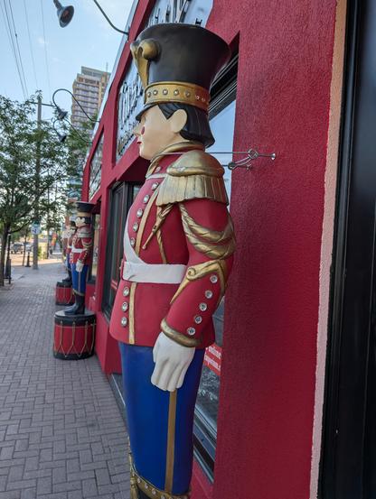 A row of life-sized toy soldiers on the sidewalk along a storefront. Each has a metal cable around its neck, presumably to keep it from tipping.
