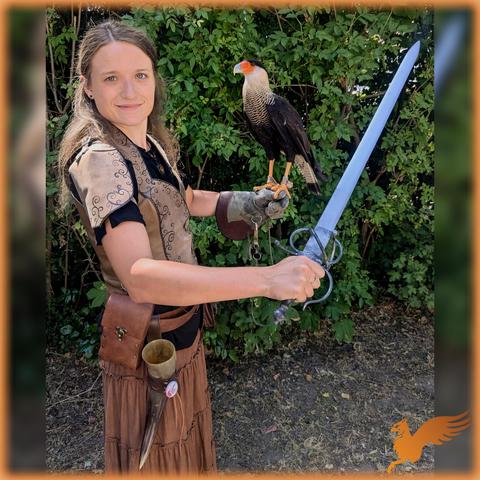 Foto auf einem Mittelaltermarkt. Ich (Frau Ende 20 mit langen, dunkelblonden Haaren) stehe in einem Fantasy-Mittelaltergewand seitlich vor der Kamera. In der rechten, vorderen Hand halte ich ein kurzes, rapierähnliches Schwert in die Höhe, auf der linken Hand sitzt ein Greifvogel mit dunklem Gefieder, weiß gesprenkelter Halskrause und rot am Kopf. Ich schaue in die Kamera, der Vogel zu mir.