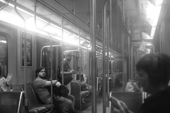 The interior of a subway car, shot through one of the acrylic barriers separating seats from the doors. Lines converge on the vanishing point at the far end of the train. We see riders standing, sitting, reading their phones. One looks at the camera, holding a backpack in his lap. Everything has a hazy, almost smoky look to it, caused by the wear to the acrylic panel between the camera and the subjects.
