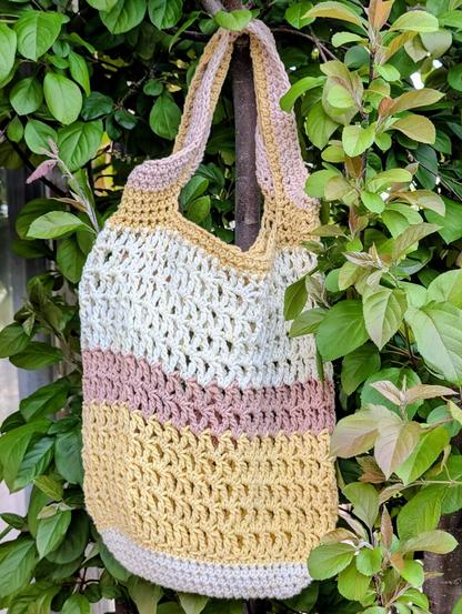 A crochet market bag in soft shades of pink, yellow, and cream tastefully hanging amongst the leaves of a crab apple tree.