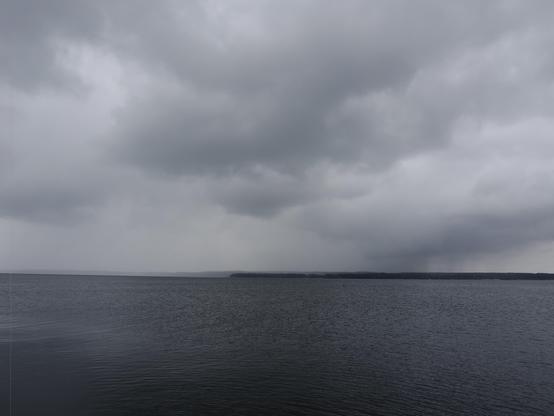 Overcast sky with thick gray clouds covering the horizon. A vast expanse of dark, calm water stretches towards a distant, barely visible coastline. The overall atmosphere is somber and dramatic.