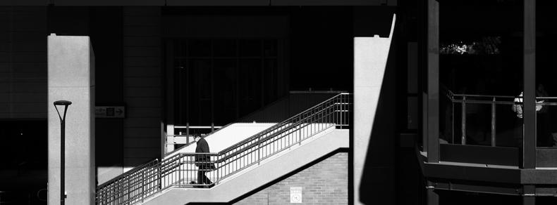 A person in dark clothing ascends an outdoor staircase with metal railings, cast in contrasting shadows and sunlight. The staircase is set against a modern building with large rectangular structures. To the right, an elevated walkway is partially visible, with another person standing near the railing. The entire scene is in black and white, emphasizing the play of light and shadow.