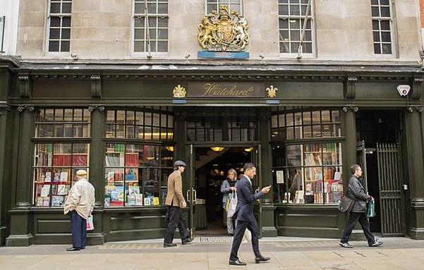 The image depicts the exterior of a bookstore named "Hatchards," which is situated on a city street. The building has a classic architectural style with a dark green facade and large windows displaying various books. Above the entrance, there is a prominent coat of arms and the name "Hatchards" in gold lettering. The windows are filled with colorful book covers, suggesting a wide selection of literature. The entrance is open, and a few people are visible in front of the store, including a man in a suit looking at his phone, a woman with a shopping bag, and an older man in a beige jacket and cap. The street is paved with light-colored stones, and the overall atmosphere is that of a busy urban environment.

Provided by @altbot, generated privately and locally using Ovis2-8B
