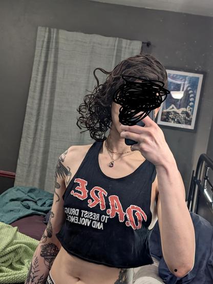 This enby taking a mirror selfie. Their hair is curly, bouncy, and fresh. They have their non-binary pendant and some colorful beads around their neck Their arm tattoos are visible including the cute frog by the bicep and the jellyshroom on the inner arm. They're wearing a black cutoff crop top which reads "D.A.R.E. to resist drugs and violence". The bottom hem of the cutoff is curled wide and upward. The toned midriff is exposed and you can see the curves of the waist into the hips along with their torso tattoo. The only other thing they're wearing is tomboyx boyshorts with glow-in-the-dark planets and asteroids.