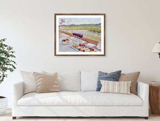 A framed landscape painting with wine, cheese, fruit, hangs on a wall. Below the painting is a couch with pillows.