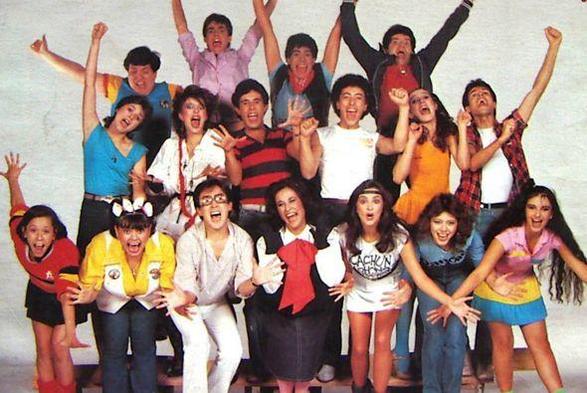 Grupo de jóvenes y adultos posando enérgicamente para la cámara, vestidos con atuendos coloridos y expresando alegría en un ambiente divertido, posiblemente un set de filmación o espectáculo. Ellos son los actores de la serie de comedia Los Cachunes de los años 80.