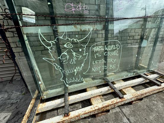 A graffiti drawing of a monster's head on a sheet of glass leaning against a building. The building is a window repair shop, so the glass is probably going into a building somewhere, but someone tagged it while it was sitting on the sidewalk. The monster has horns and is saying, "The world is cruel. Let's try not to be."