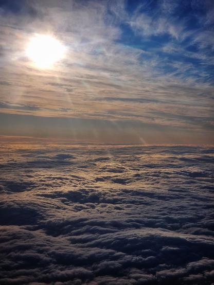 An aerial view from an aeroplane shows a vast expanse of fluffy white clouds stretching to the horizon, illuminated by a bright, warm sun on the left side of the frame. The sky above the clouds transitions from a soft orange and pink near the horizon to a deeper blue with wispy white cirrus clouds higher up. The clouds below are densely packed, resembling a textured blanket, with some areas catching the golden light of the setting or rising sun, creating a serene and expansive atmosphere.