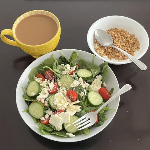 A simple salad of arugula, spinach, grape tomatoes, cucumber slices, crumbled goat cheese, and extra virgin olive oil drizzled over the salad and seasoned with salt and pepper. On the side, there’s a bowl of vanilla yogurt with granola on top and a cup of almond coffee