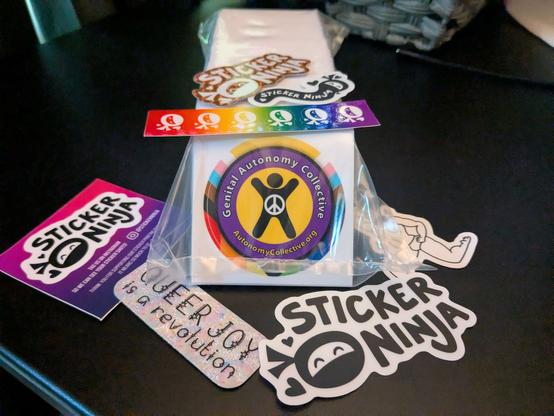 A stack of Genital Autonomy Collective Logo stickers wrapped in plastic surrounded by complimentary Sticker Ninja stickers including one that says QUEER JOY is a revolution.