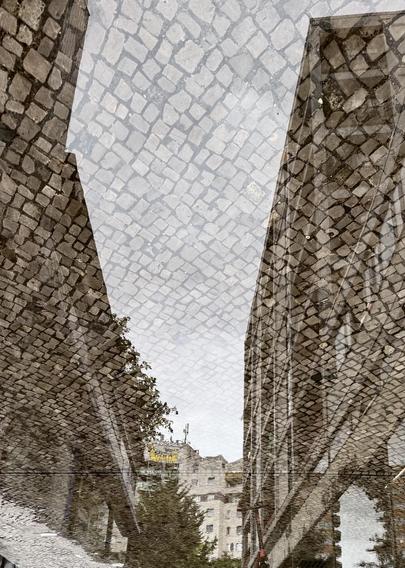 A reflection of cobblestone streets in a puddle, with buildings visible on either side under a cloudy sky.
