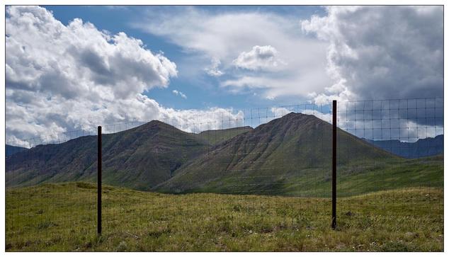 Green grassy hillside with a metal wire fence in the foreground and rolling hills under a partly cloudy sky in the background. Two fence posts are prominent; the left post is taller, standing isolated against the sky.   The fence itself is wire mesh with large holes (a wildlife fence)