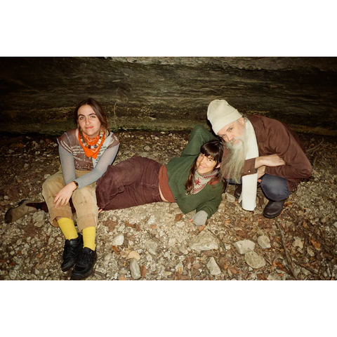 Three people look towards the camera. They appear to be on the floor of a cave and the flash makes the photo slightly overexposed. The person on the left is white with long brown hair, an orange scarf, yellow pants, and a denim jacket. The person in the middle is white with long dark hair, a green sweater, and brown pants. The person on the right is wearing a white beanie, brown jacket, and has a long white beard.