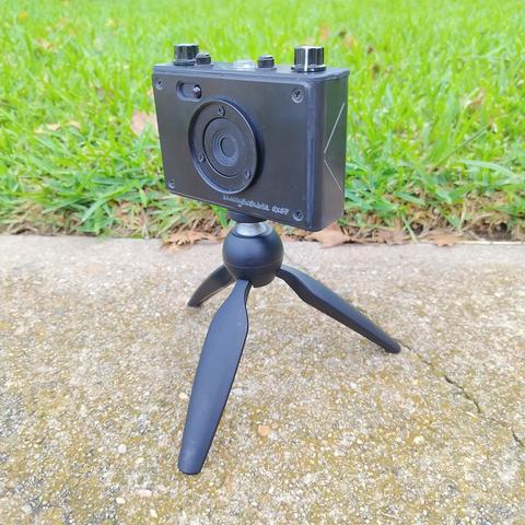 A RealitySoSubtle 6x6F pinhole camera mounted to a small Manfrotto desktop tripod, sitting on concrete in front of some green grass.