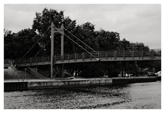 Auf dem Bild sieht man die Fußgänger- und Fahrradbrücke, die über die Peene in Anklam führt. Diese kleine Hänge- oder Schrägseilbrücke verbindet das Stadtzentrum mit dem Uferbereich und dem Hafen. Die Brücke ist aus Holz und Metall konstruiert und hat einen ganz eigenen Charme — besonders in Schwarz-Weiß wirkt sie fast nostalgisch.