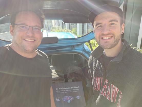Auf dem Bild sind zwei männlich gelesene Personen zu sehen, links Michael Pelzer, rechts David Mayer, die in die Kamera lächeln. Im Hintergrund ist der offene Kofferraum eines Autos zu sehen und die Spielebox des KI-Escape-Games.