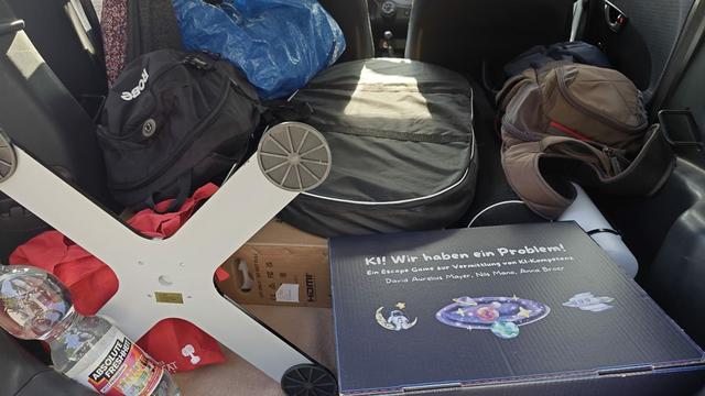 Auf dem Bild ist ein voll beladener Kofferraum zu sehen. Unter anderem sieht man Rucksäcke, Kartons, einen Tischfuß und eine Spielbox, auf der steht "KI! Wir haben ein Problem! Ein Escape Game zur Vermittlung von KI-Kompetenz. David Aurelius Mayer, Nils Mano, Anna Broer".