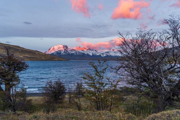 Sunrise view of the Torres del Paine spires from the shores of Laguna Azul in Chilean Patagonia. The peaks are dramatically lit in deep red by the first sunlight, while the landscape below remains in cool, shadowed tones near the blue lagoon.